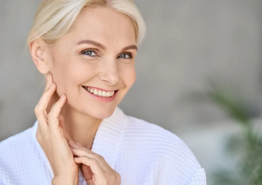 A woman with radiant skin smiling and touching her neck to show the results of treatment as part of her neck filler aftercare.