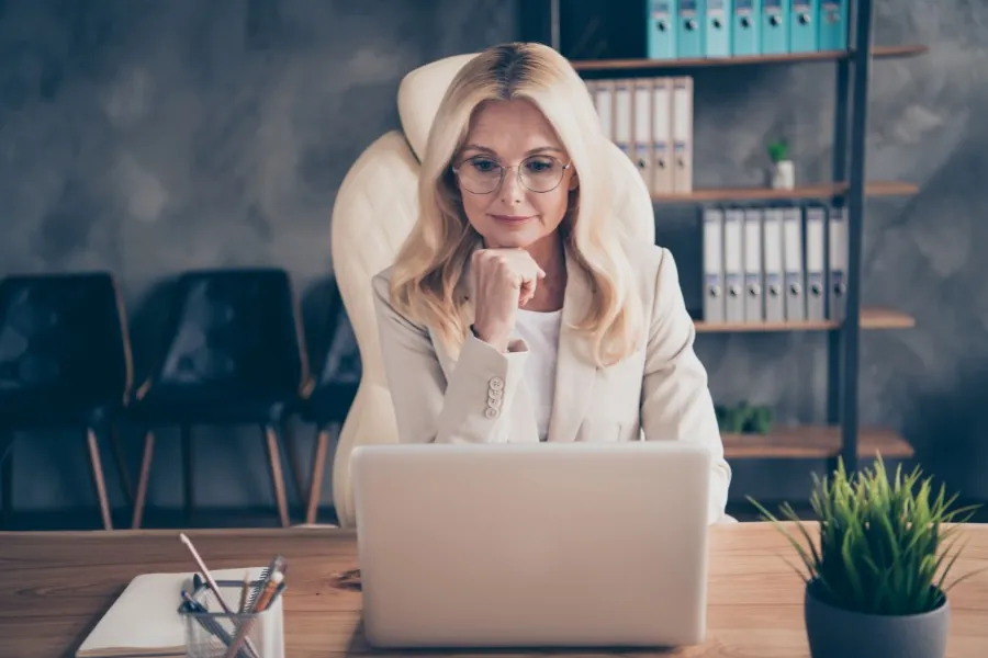 Blonde woman with glasses sitting at desk using laptop with neutral expression